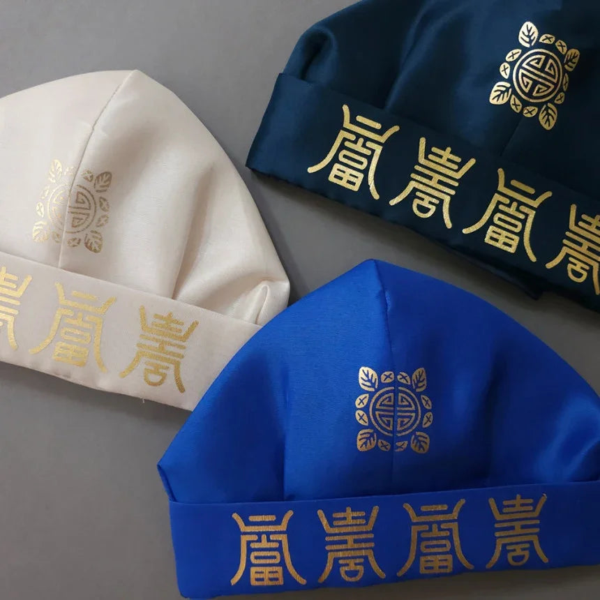 Three Yuhan Gunmo traditional Korean hats in white, navy, and blue with gold embroidery by Native Korean