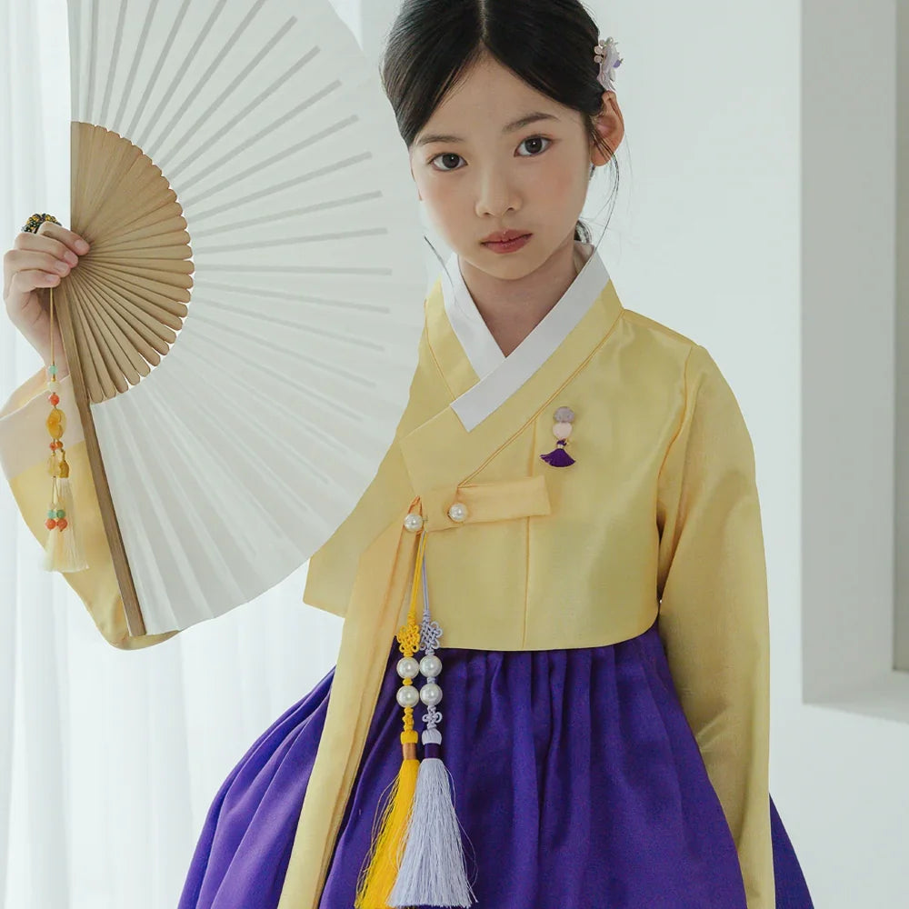 Girl wearing a yellow and purple Korean hanbok holding a white folding fan.