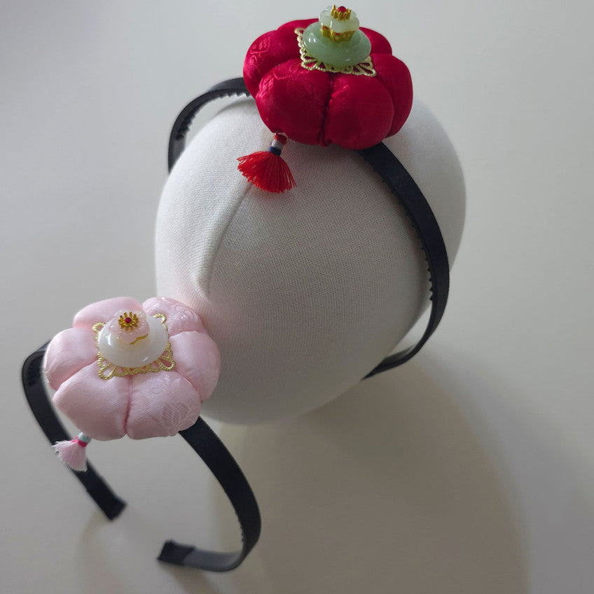 A decorative black headband displayed on a white mannequin head, featuring two fabric flower ornaments: one in bright red with a small green and gold accent, and one in pale pink with a white and gold accent.